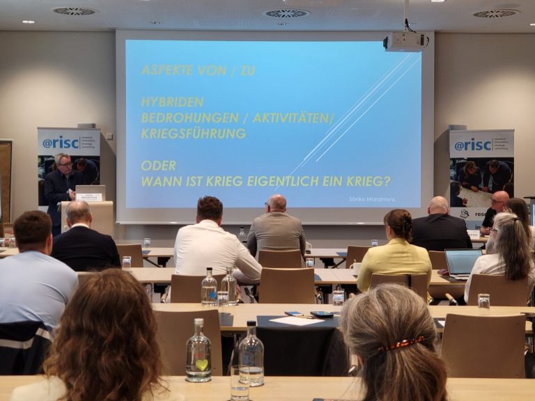 Konferenzraum mit Referent und Publikum, Präsentation von Sönke Marahrens über hybride Bedrohungen.